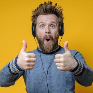 Ein freudig überraschter Mann mit Kopfhörern hört Musik und macht eine anerkennende Geste, den Daumen hoch. Gelber Hintergrund.